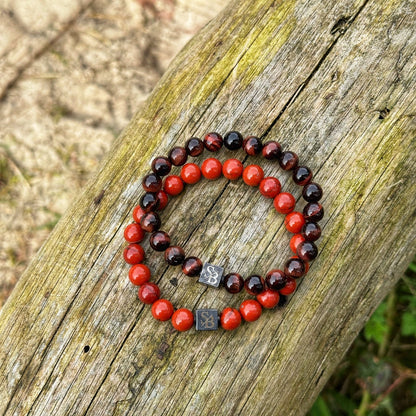 Twee kralen armbanden van Stoney Bracelets liggen op een verweerd houten oppervlak. De ene armband is voorzien van donkerbruine kralen, terwijl de andere felrode kralen van Red Sesame Stones (8mm) toont. Beide armbanden hebben vierkante metalen bedels met een gegraveerd ontwerp. De achtergrond toont vuil en wat groen, wat de rijke natuursteen-aantrekkingskracht van de scène versterkt.