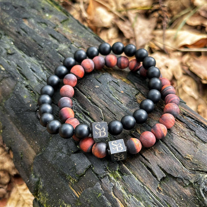 Twee kralen armbanden rusten op een stuk verweerd hout. De ene armband heeft zwarte kralen, terwijl de andere rode houten kralen heeft. Beide armbanden, genaamd Frosted Tiger Eye Stones (8mm) van Stoney Bracelets, hebben een klein vierkant bedeltje met de initialen "SB" erin gegraveerd. Op de achtergrond zijn gevallen bladeren zichtbaar.