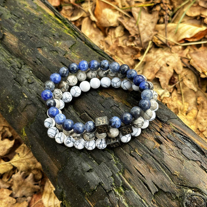 Twee kralenarmbanden van Stoney Bracelets, met 8 mm Lapis Lazuli-stenen afgewisseld met witte en grijze natuursteenstenen, rusten op een verweerde boomstam tussen gevallen bladeren. Elke armband is versierd met een klein vierkant bedeltje gegraveerd met een gouden "SB". Het tafereel lijkt zich af te spelen in een bebost gebied.