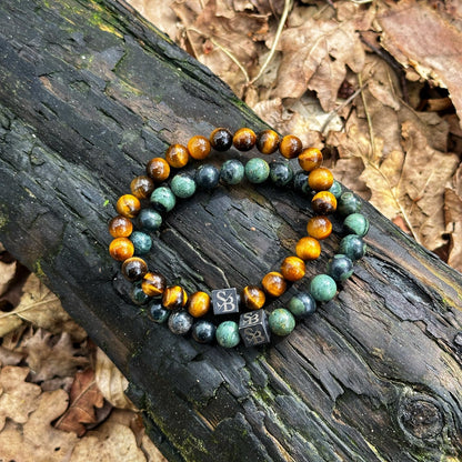 Twee kralen armbanden van Stoney Bracelets liggen op een donkere, verweerde boomstam te midden van een achtergrond van droge bladeren. De ene armband heeft afwisselend Gold Tiger Eye Stones (8mm) en groene edelsteen kralen, terwijl de andere voornamelijk bestaat uit groene natuursteen kralen. Beide armbanden hebben kleine, vierkante metalen bedels met een gegraveerd symbool.