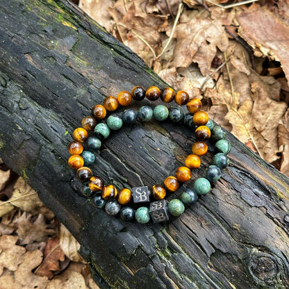 Twee elastische kralenarmbanden van Stoney Bracelets rusten op een met mos bedekte boomstam, omringd door gevallen bruine bladeren. Eén armband, met de Kambaba Jasper Stones (8mm), toont kralen in groene en blauwe tinten, terwijl de andere glanzende goudbruine kralen toont. Beide armbanden zijn versierd met kleine, vierkante metalen bedels.