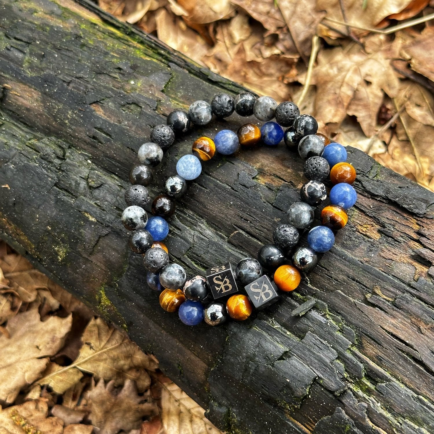 Twee kralenarmbanden uit de Rainforest | Mixed Stones (8mm) collectie van Stoney Bracelets zijn op een verweerde boomstam geplaatst. Elk is voorzien van een mix van zwarte, blauwe en bruine kralen, waarbij er één een klein vierkant bedeltje toont met de gravure "SB". De boomstam rust op de grond bedekt met bruine, gedroogde bladeren.