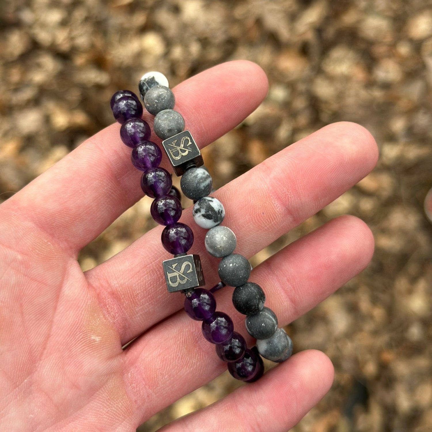 Een hand houdt twee armbanden vast uit de Stoney Bracelets-collectie. De ene, versierd met paarse transparante kralen, vult de andere aan, die is gemaakt van Frosted Zebra Stones (8 mm). Beide hebben een klein vierkant metalen bedeltje met een gegraveerd ontwerp. De achtergrond is wazig en lijkt buiten te zijn.