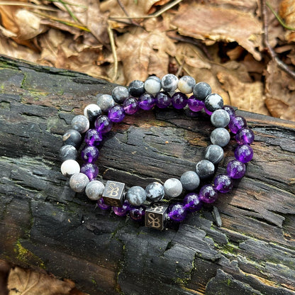 Twee kralen armbanden van Stoney Bracelets liggen op een donkere, verweerde boomstam, omringd door droge bladeren. De ene armband is voorzien van zwart-witte gemarmerde natuurstenen kralen, terwijl de andere 8mm Amethist Stenen toont. Beide armbanden zijn versierd met een kleine metalen vierkante bedel met een symbool erin gegraveerd.