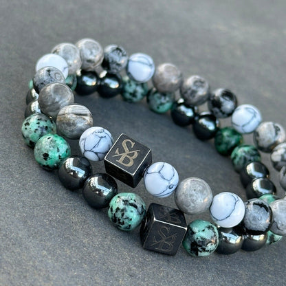 Een paar Stoney Bracelets op een grijs oppervlak. De armbanden, genaamd Natural | Mixed Stones (8mm), bevatten een mix van edelstenen in tinten blauw, groen en wit, samen met enkele zwarte kralen. Elke armband bevat een zwarte kubuskraal met een gegraveerd ontwerp.