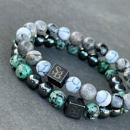 Twee Stoney Bracelets "Nautic | Mixed Stones (8mm)" armbanden worden op een grijs oppervlak getoond. Elk heeft een mix van gemarmerde witte, grijze, groene en zwarte natuursteen kralen. Beide hebben een kubusvormige kraal met de letters "SB" erin gegraveerd. De kralen variëren in textuur en patroon.