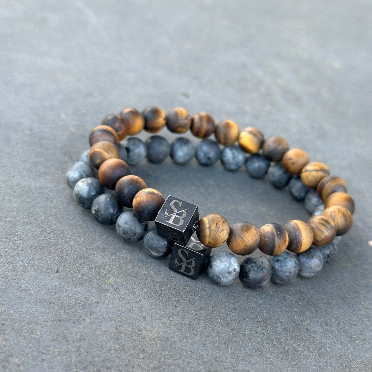 Twee kralenarmbanden rusten op een plat oppervlak. De ene is voorzien van donkerbruine Frosted Gold Tiger Eye Stones (8mm) van Stoney Bracelets met strepen, terwijl de andere grijs gemarmerde natuursteen toont. Beide armbanden hebben een kubieke bedel met gegraveerde letters.