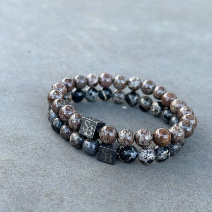 Twee elastische armbanden van Stoney Bracelets gemaakt van ronde, gepolijste Coffee Snowflake Stones (8mm). De ene armband benadrukt bruine en grijze stenen, terwijl de andere donkergrijze en zwarte stenen laat zien. Elke armband heeft een kleine vierkante kraal met een gegraveerd embleem. Ze zijn geplaatst tegen een effen grijze achtergrond, waardoor de natuurlijke schoonheid van de edelsteen wordt geaccentueerd.