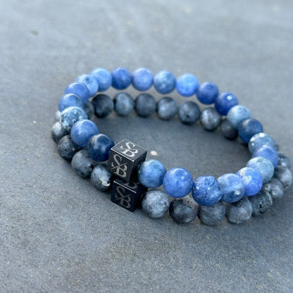 De Matte Lapis Lazuli Set (8mm) van Stoney Bracelets, met een paar kralenarmbanden met blauwe en grijze kralen, is op een plat oppervlak geplaatst. Elke armband bevat een zwarte kubusvormige kraal met unieke zilveren gravures.