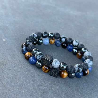 Een paar Dark Polar | Mixed Stones (8mm) kralenarmbanden van Stoney Bracelets worden op een grijs oppervlak getoond. De armbanden hebben een combinatie van zwarte, blauwe en bruine kralen met verschillende texturen en patronen. De zwarte kralen zijn gemaakt van natuursteen en lijken kleine logo's te hebben die erop zijn gegraveerd.