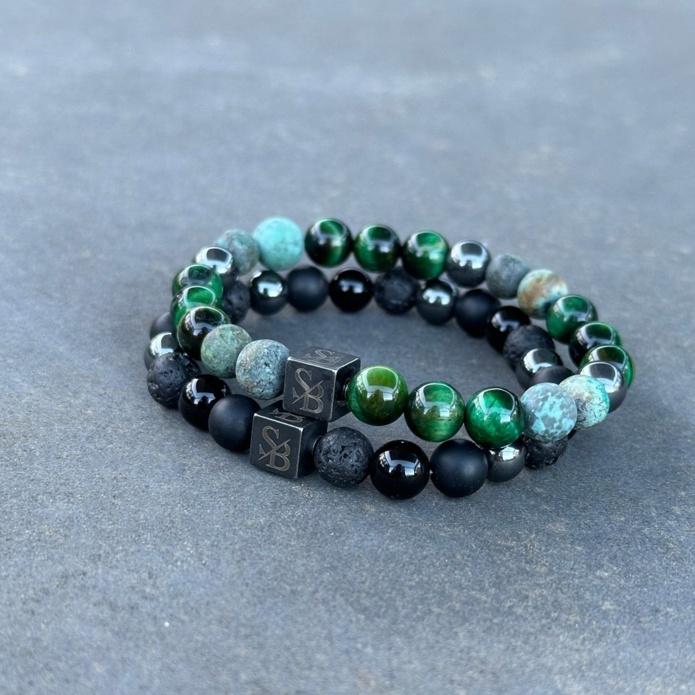 Twee Stoney Bracelets uit de Prehistoric | Mixed Stones (8mm) collectie liggen op een grijs oppervlak. Eén armband heeft een combinatie van groene en zwarte kralen met twee kubusvormige zwarte kralen met de markering "SB". De tweede armband heeft een mix van turquoise en zwarte edelsteen kralen. Beide armbanden hebben een rustieke, geparelde uitstraling.