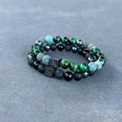 Een paar Swamp | Mixed Stones (8mm) kralenarmbanden van Stoney Bracelets worden getoond tegen een grijze achtergrond. De armbanden hebben een combinatie van groene, zwarte en steenkleurige kralen, waaronder kubusvormige kralen met symbolen erop. Gemaakt met natuursteen en edelsteen kralen, zijn de armbanden opgerold en liggen ze naast elkaar.
