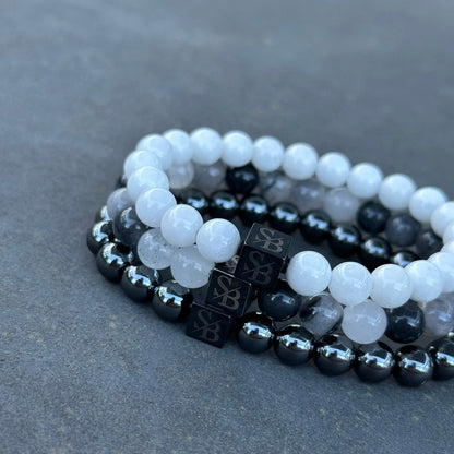 Drie kralenarmbanden uit de Angel Set (8mm) van Stoney Bracelets zijn naast elkaar geplaatst op een donkergrijs oppervlak. De armbanden bestaan uit witte, grijze en metallic zwarte kralen. Elke armband heeft een zwarte kubusvormige kraal met een logo erop, met natuursteen voor een vleugje elegantie.