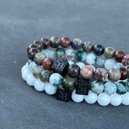 Drie armbanden uit de Apollo Set (8mm) van Stoney Bracelets op een grijs oppervlak. Elk heeft een mix van glanzende, veelkleurige ronde kralen en zwarte vierkante kralen geëtst met een ontwerp. De bovenste armband heeft aardse natuursteen kralen; de middelste heeft een vergelijkbaar patroon, terwijl de onderste witte edelsteen kralen toont.