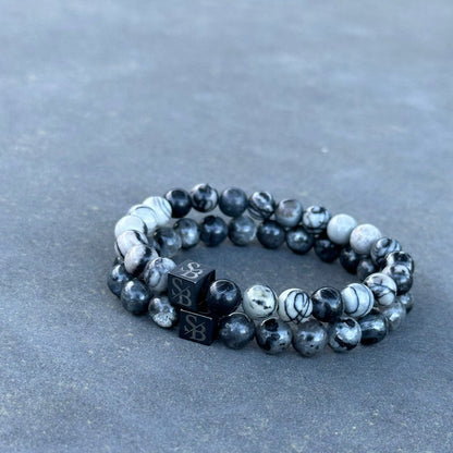 Een paar armbanden van Stoney Bracelets, bekend als Black Silk Stones (8mm), zijn gemaakt van grijs en zwart gemarmerd natuursteen en gerangschikt in een cirkel. Elke armband heeft een zwarte kubieke kraal met zilveren letters "S" en "B" erin gegraveerd. De achtergrond is een grijs, gestructureerd oppervlak.