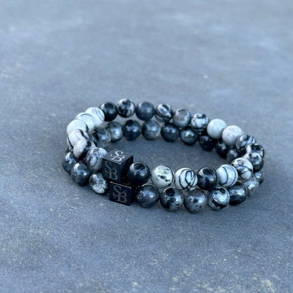 Een paar gemarmerde zwart-witte Double Gray Set (8mm) armbanden van Stoney Bracelets liggen op een glad grijs oppervlak. Elke armband heeft een zwarte kubuskraal gegraveerd met de letters "SB", wat een contrasterend ontwerp toevoegt aan het algehele uiterlijk.