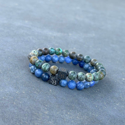 Twee kralenarmbanden van de Seafloor Set (8mm) van Stoney Bracelets worden op een plat oppervlak gelegd. Elke armband bestaat uit ronde kralen in verschillende kleuren; de ene heeft voornamelijk groenblauwe kralen, terwijl de andere blauwe kralen heeft. Beide hebben zwarte kubieke kralen gegraveerd met een "SB"-logo.