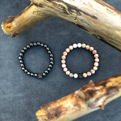 Twee armbanden zijn op een plat, donker oppervlak geplaatst. Degene aan de linkerkant is gemaakt van glanzende, donkere Hematiet Stones (8mm) van Stoney Bracelets, terwijl degene aan de rechterkant bestaat uit gladde, lichtgekleurde kralen. Twee houten takken zijn gedeeltelijk zichtbaar in de linkerbovenhoek en de rechteronderhoek.