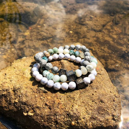 Een drietal kralenarmbanden uit de Triple White Set (8 mm) van Stoney Bracelets, met een mix van grijze, witte en groene stenen, zijn op een grote rots bij een waterpartij geplaatst.