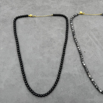 Twee kettingen rusten op een grijs oppervlak: de Stoney Bracelets Matte Obsidian Necklace (6mm) aan de linkerkant heeft ronde zwarte kralen voor kracht en bescherming, terwijl de rechter ketting een mix heeft van donkere en lichte gespikkelde vulkanische steen kralen. Beide kettingen worden geleverd met goudkleurige sluitingen en kettingen.