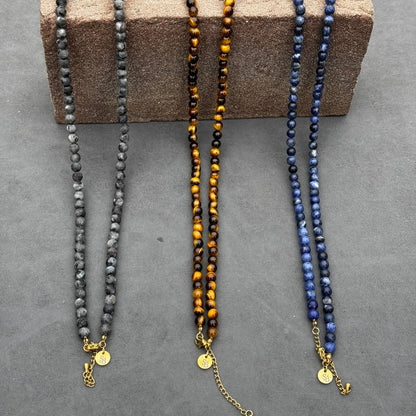 Drie Royal Lapis Necklaces (6mm) van Stoney Bracelets, elk met gouden sluitingen en kleine ronde bedels, worden op een grijs oppervlak getoond. Hun kralen in tinten grijs, bruin en blauw rusten op een getextureerd bruin blok.