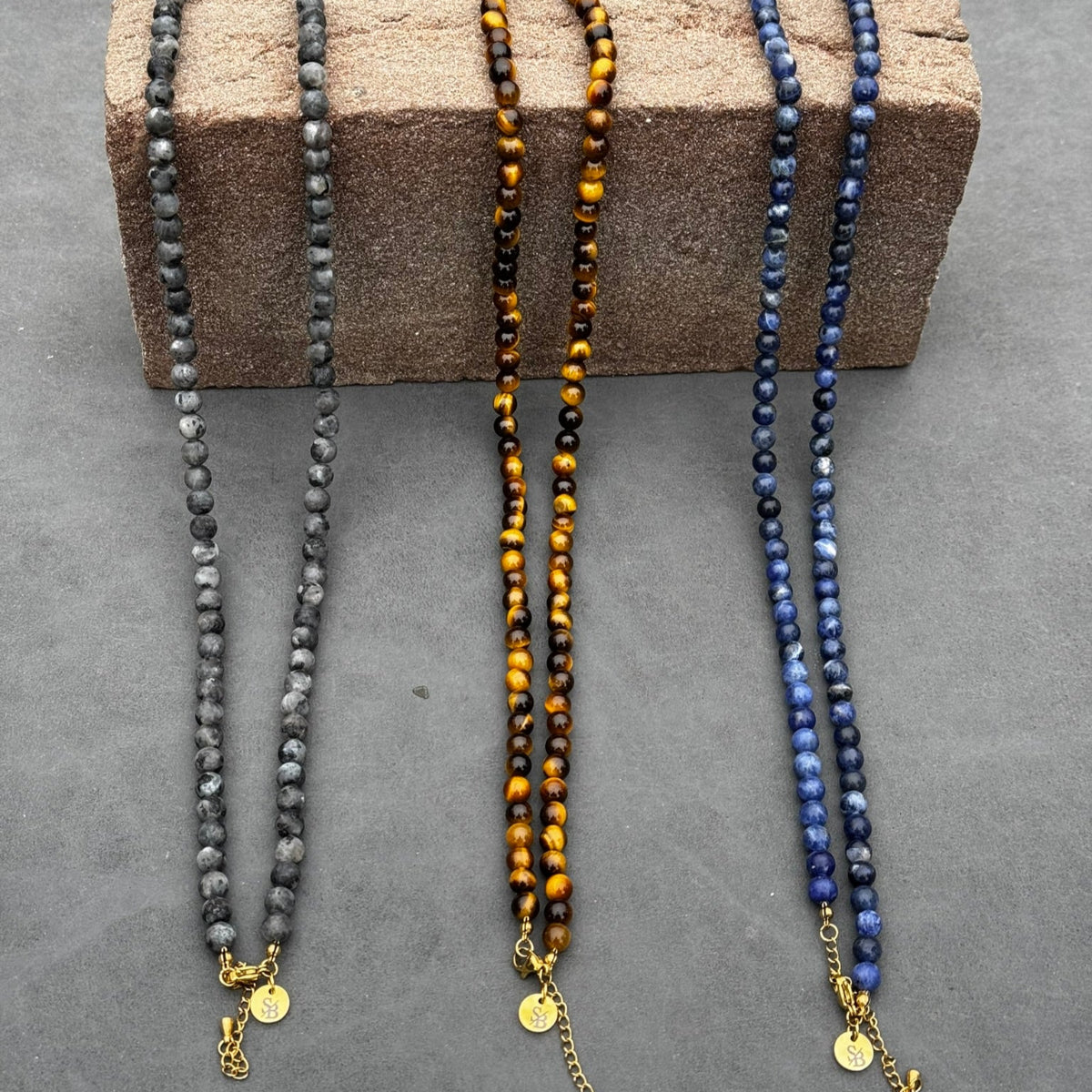 Drie Golden Gaze Necklaces van Stoney Bracelets worden getoond op een getextureerd blok tegen een grijze achtergrond. De 6mm kralen zijn verkrijgbaar in grijs, blauw en tijgeroog tinten. Elk heeft een klein gouden bedeltje en een kettingverlenging.