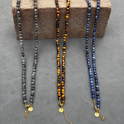 Drie kralenkettingen van Stoney Bracelets worden op een getextureerd bruin blok getoond. De linkerketting is de Matte Moon Necklace (6 mm) in grijs, die emotionele balans bevordert, de middelste heeft tijgeroog en de rechter is blauw. Elk heeft een gouden sluiting en een ronde bedel.