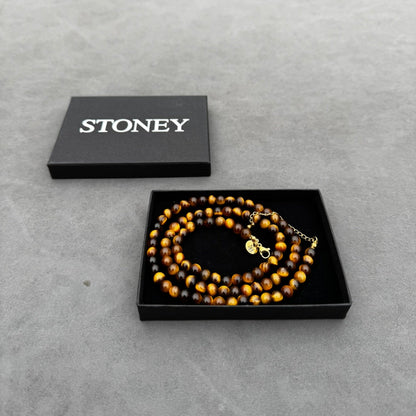 Een zwarte Stoney Bracelets doos is open en onthult de Royal Lapis Necklace (6mm) gemaakt van afwisselend bruine en zwarte kralen met een klein gouden bedeltje. De elegante ketting rust sierlijk op een grijs oppervlak.