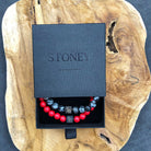 Een zwarte doos met het label "Stoney Bracelets" bevat de Red vs Black Set (8mm), met twee natuursteen kralenarmbanden. De ene heeft rode kralen met zwarte afstandhouders en de andere heeft zwarte kralen met witte marmering. De doos is geplaatst op een plak gepolijst hout met zichtbare nerfpatronen.