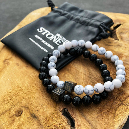 Twee kralen armbanden, een met Obsidian Stones (8mm) van Stoney Bracelets en de andere met witte gemarmerde natuurstenen, liggen op een houten oppervlak. Elke armband is versierd met een vierkante metalen kraal gegraveerd met de initialen "SB." Een zwart trekkoordzakje met het label "Stones" rust op de achtergrond.