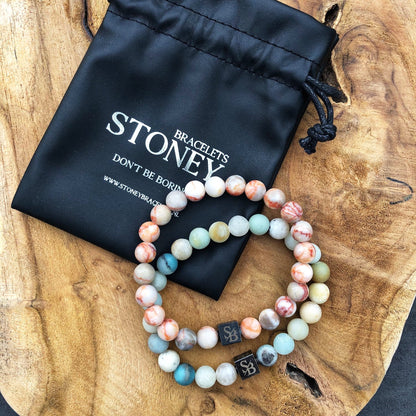 Twee kralenarmbanden met levendige Marsstenen (8 mm) rusten op een houten oppervlak naast een zwart trekkoordzakje met "Stoney Bracelets" erop gedrukt. Het zakje toont ook de slogan "DON'T BE BORING" en een website-URL.