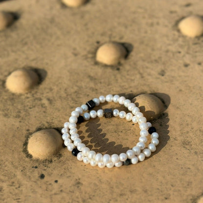 De Rounded Pearl Bracelet I (7mm) van Stoney Bracelets ligt op een gestructureerd natuursteen oppervlak met grote ronde bulten. De armband bestaat uit witte parels met een paar zwarte kralen ertussen en een kleine metalen sluiting. Het oppervlak lijkt verweerd en gevlekt.