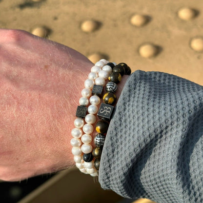 Een hand is versierd met meerdere Stoney Bracelets, waaronder de Rounded Pearl Bracelet I (7mm). De armbanden zijn voorzien van witte parelkralen, kleine zwarte kralen en kralen met zilveren accenten. De persoon draagt een grijs shirt met lange mouwen en textuur. De achtergrond is licht vervaagd met enkele ronde objecten zichtbaar.