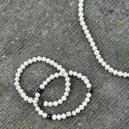 Close-up van een parelsieraad op een getextureerd, grijs oppervlak. De set bestaat uit een parelketting en twee Stoney Bracelets' Rounded Pearl Bracelet I (7mm). De armbanden zijn versierd met kleine zwarte en zilveren decoratieve kralen die tussen de parels zijn verspreid, wat een vleugje elegantie toevoegt met kralen en natuursteenelementen.