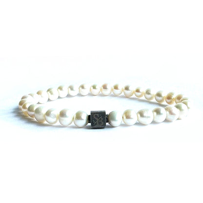 Introductie van de Rounded Pearl Bracelet I (7mm) van Stoney Bracelets: Een ronde armband met gelijkmatig verdeelde witte parels, aangevuld met een kleine, vierkante metalen sluiting met gegraveerde initialen. Het elegante stuk is gefotografeerd tegen een effen, witte achtergrond.