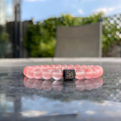 Een Stoney Bracelets armband met 6mm Cherry Quartz Stones en een enkele, vierkante zwarte natuursteen kraal met een gouden design rust op een reflecterend oppervlak. De achtergrond toont wazig groen en een deel van een structuur onder een blauwe lucht met een paar wolken.