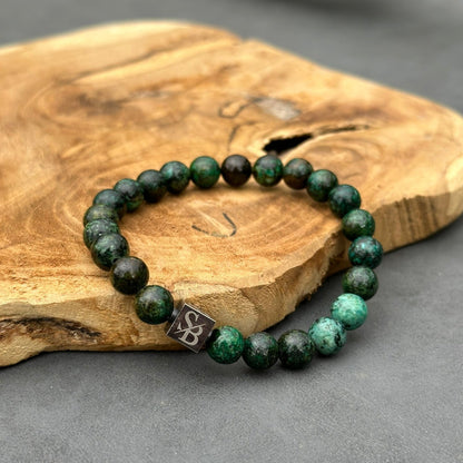 Een kralenarmband gemaakt van Stoney Bracelets' Dark African Turquoise Stones (8mm) in donkergroen en zwart is geplaatst op een stuk glad, lichtbruin hout. De armband heeft een metalen vierkante bedel met een gegraveerd ontwerp. De achtergrond is een grijsachtig oppervlak.