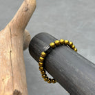 Een Stoney Bracelets-stuk, de Gold Hematiet Stones (8mm) armband, met een zwarte rechthoekige bedel gegraveerd met de initialen "SB", wordt elegant tentoongesteld op een donker cilindrisch object. Een stukje drijfhout voegt een natuurlijke toets toe aan de scène. De neutrale grijze achtergrond versterkt de verfijnde aantrekkingskracht van de kralen in dit elegante accessoire.