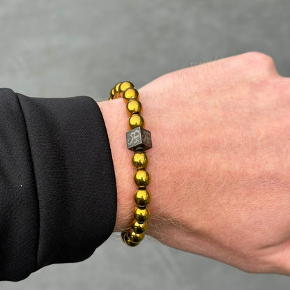 Een persoon draagt een "Stoney Bracelets" armband gemaakt van 8mm Gold Hematite Stones, inclusief een zwarte kubusvormige kraal met een ontwerp. De persoon draagt een zwarte mouw en de achtergrond is een effen grijs oppervlak.