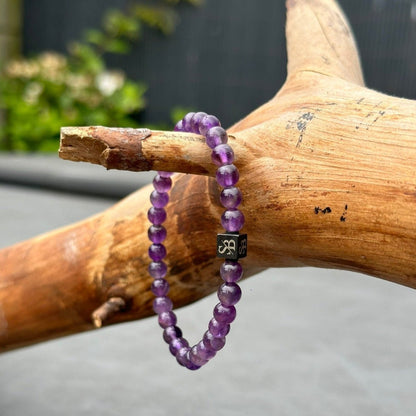 Een Stoney Bracelets-creatie met Amethiststenen (6 mm) en een kleine zwarte kubus versierd met een wit ontwerp, mogelijk gemaakt van natuursteen, is elegant gedrapeerd over een houten stok. De achtergrond toont wazig groen gebladerte en een grijs oppervlak.