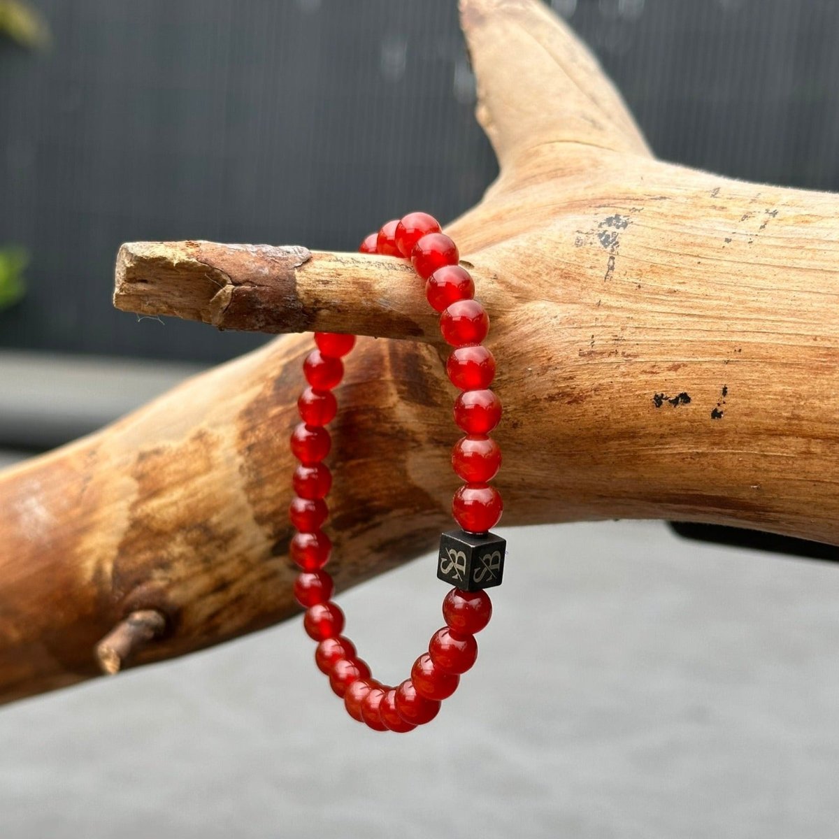 Een *Stoney Bracelets* Red Onyx Stones (6mm) armband, versierd met een centrale zwarte vierkante bedel met een symbool, wordt getoond op een getextureerde houten tak tegen een neutrale achtergrond. De kralen zijn gelijkmatig rond en gepolijst.