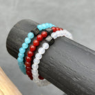 Drie kralenarmbanden van Stoney Bracelets worden op een donkere houten cilinder tegen een grijze achtergrond getoond. Eén armband toont blauwe kralen, een andere heeft rode kralen en de derde is versierd met de White Onyx Stones (6 mm). Elke armband bevat een zwarte kubusvormige kraal met een ingewikkeld ontwerp.