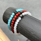 Drie kralenarmbanden uit de Aquamarine Stones (6mm) collectie van Stoney Bracelets worden tentoongesteld op een donker cilindrisch object. De armbanden, met respectievelijk turquoise, rode en witte natuursteen kralen, tonen elk een kleine zwarte kubus met een bedrukt ontwerp. De achtergrond is een effen grijs oppervlak.