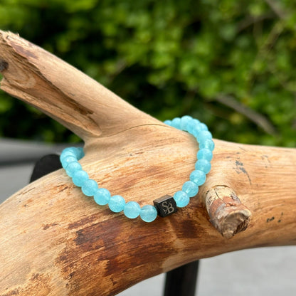 Een Aquamarine Stones (6mm) armband van Stoney Bracelets, met een lichtblauw kralenontwerp en een zwarte vierkante bedel met een ingewikkeld patroon, is gedrapeerd over een verweerd, lichtbruin stuk drijfhout. De achtergrond toont wazig groen gebladerte.