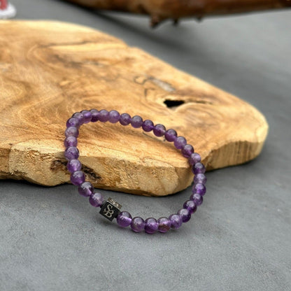 Een kralenarmband gemaakt met Stoney Bracelets' Amethyst Stones (6mm) is geplaatst op een getextureerd houten oppervlak. Er is een enkele zwarte vierkante kraal met een ontwerp erop. De achtergrond is een grijs oppervlak.