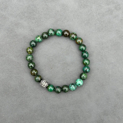 Een kralenarmband van Stoney Bracelets met 8 mm donkere Afrikaanse turquoise stenen in een cirkelvormig patroon tegen een grijze achtergrond. Eén van de stenen is versierd met een klein vierkant, metalen bedeltje met de tekst 'SB' erin gegraveerd.