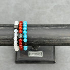 Drie kralenarmbanden tentoongesteld op een zwarte standaard. De armbanden bevatten er een met witte onyxstenen (8 mm), een met rode kralen en een met blauwe kralen. Elke armband heeft een kleine vierkante metalen kraal met een decoratief ontwerp. De standaard heeft een plaquette met het label "Stoney Bracelets.