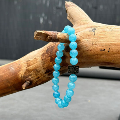 Een armband van Stoney Bracelets genaamd Aquamarine Stones (8mm), met gepolijste, ronde, lichtblauwe kralen, is elegant gedrapeerd over een stuk natuurlijk hout. De armband bevat een zwarte vierkante bedel versierd met een zilveren design. De achtergrond is wazig, waardoor de armband en het hout worden geaccentueerd.