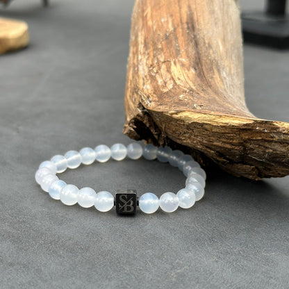 Een witte kralenarmband met witte onyxstenen (8 mm) en een zwarte vierkante bedel met de letters "SB" van Stoney Bracelets liggen op een donkergrijs oppervlak naast een groot, gebogen stuk natuursteen.