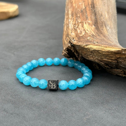 Een opvallende armband van Stoney Bracelets met ronde, blauwe aquamarijnstenen van 8 mm en een opvallende, enkele, zwarte kubieke kraal met de initialen "SB" erin gegraveerd. De armband wordt elegant tentoongesteld op een donker oppervlak naast een stuk verweerd hout, waardoor de charme van natuursteen perfect tot zijn recht komt.